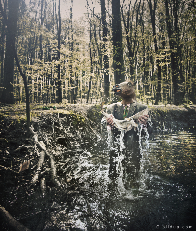 Knaldemand is standing in a river in a forest with the sun shining on him through the leaves. He is wearing a suit and holding a cod that he just caught.